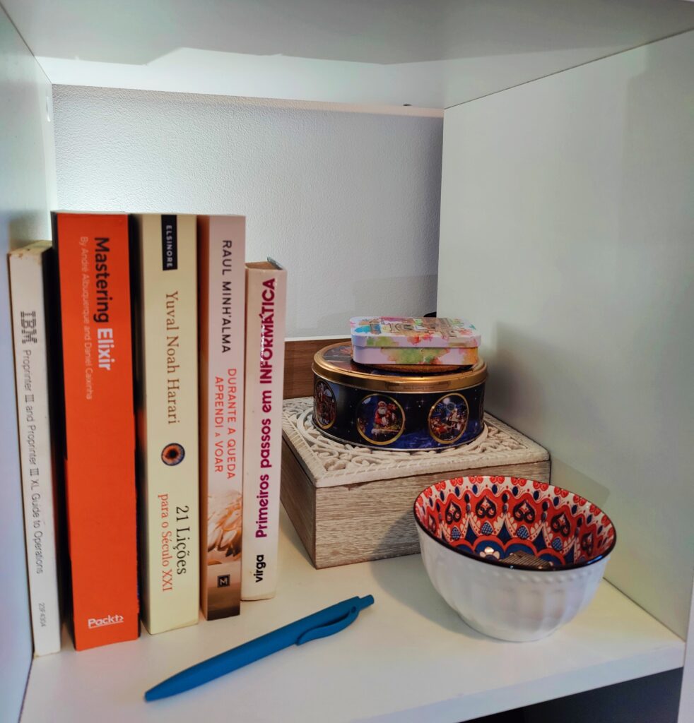 A shelf with books on the left, a tin cookie box on the right atop a small box, a pen and bowl are also visible.